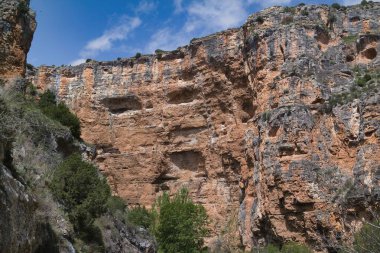 Büyük kayalıklar belirgin bir şekilde yükseliyor, kayalık dokular ve toprak renkler sergiliyor. Yeşillik üste gelişir, engebeli oluşumun aksine. Açık mavi gökyüzü sahneyi büyütüyor.