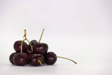 Koyu kırmızı kirazlar pürüzsüz beyaz bir yüzey üzerinde bir araya toplanarak parlak ciltlerini ve ince gövdelerini vurguluyor ve çekici bir natürmort oluşturuyorlar..