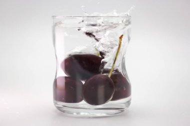 Kirazlar suyla dolu berrak bir bardağa düşer, canlı su sıçramalarına ve dalgalanmalara neden olur. Fotoğraf çarpışma anını yakalıyor, tazelik ve canlılık sergiliyor..