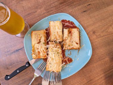 Peynirle kaplanmış ve yanında sos ile birlikte makarnanın katmanlarının bulunduğu mavi tabakta sunulan tuzlu bir yemek. Büyük bir bardak bira yakınlarda kırsal bir ahşap masada oturuyor..