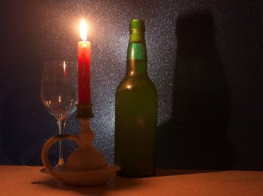 Mum ışığı bir kadeh şarap ve yeşil bir şişenin yanında yanıp sönüyor. Sıcak bir ortam yaratıyor. Beyaz bir tutucu, ayarlara sıcaklık katan canlı kırmızı bir mum sergiliyor.