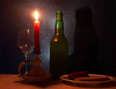 Titrek bir mum, boş bir şarap bardağı, yeşil bir şişe ve dokulu bir arka planda atıştırmalık tabağı içeren samimi bir akşam yemeği. Sıcak ışık romantik bir atmosfer yaratır..