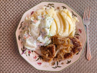 Çiçekli bir tabakta, kremalı patates püresi, sebzeli taze lahana salatası ve karamelli soğanla süslenmiş baharatlı etten oluşan doyurucu bir yemek sunulur..