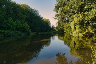 Nehrin sakin suları etrafındaki ağaçları yansıtarak sakin bir atmosfer yaratır. Güneş arka planda yükselir, yemyeşil manzarayı hafif bir ışıkla aydınlatır..