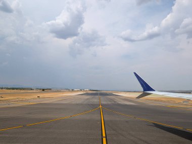 Bir uçak kalkışa hazır bir şekilde pistte bekliyor. Manzara açık gökyüzü ve bulutların karışımıyla genişliyor. Arka planda kentsel büyümeyi gösteren bir şehir silüeti görülebilir.