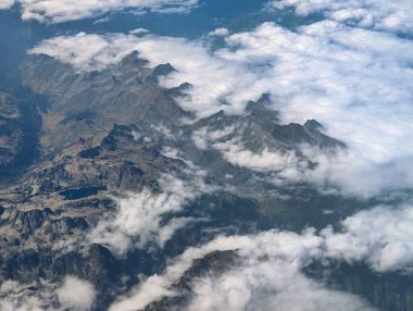 Güneşli bir günde kalın bir bulut tabakasından yükselen görkemli dağ zirvelerini görüntüleyen hava manzarası aşağıdaki büyüleyici ve engebeli manzarayı gözler önüne seriyor. Doğanın güzelliği vurgulanıyor..