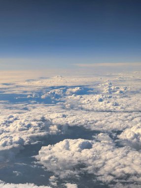 Pofuduk beyaz bulutlar, gün ışığında bir uçaktan yakalanan uçsuz bucaksız mavi gökyüzünün altındaki dünyayı kaplıyor. Sahne huzur ve merak taşır..