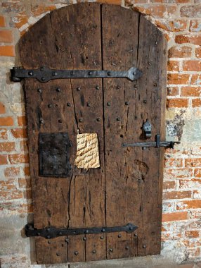 Eski kerestelerden yapılmış sağlam ahşap bir kapı, tarih ve zaman belirtileri gösteren yıpranmış tuğla bir duvarın içine yerleştirilmiş demir menteşeler ve sağlam bir mandal içerir..