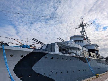 Limana yanaşan tarihi bir savaş gemisi, büyük topçu ve modern donanım gösteriyor. Bulutlu gökyüzü dramatik bir zemin oluşturuyor ve geminin tarihsel önemini vurguluyor..