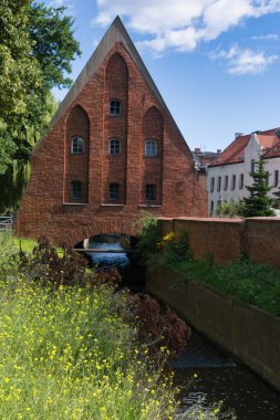 Eşsiz bir mimari tasarımı olan çarpıcı bir kırmızı tuğla yapı barışçıl bir kanalın yanında duruyor. Çevresindeki bölgede mavi gökyüzünün altında canlı yeşil çimenler ve kır çiçekleri var..