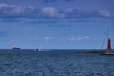 İki gemi limana yakın sakin sularda seyahat eder. Küçük bir tekne mavi gökyüzünün altında kırmızı bir fenere yaklaşırken, büyük bir kargo gemisi uzaktadır..