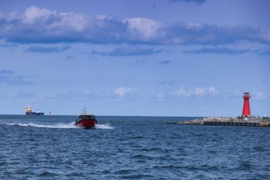 Daha büyük bir gemi mesafeye demir attığı için parlak kırmızı bir tekne durgun sularda hızla ilerliyor. Kıyı feneri kıyıda dimdik durur, kıyı manzarasını güzelleştirir..