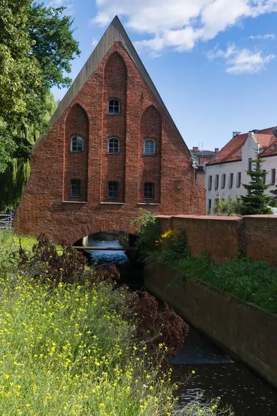 Eşsiz bir mimari tasarımı olan çarpıcı bir kırmızı tuğla yapı barışçıl bir kanalın yanında duruyor. Çevresindeki bölgede mavi gökyüzünün altında canlı yeşil çimenler ve kır çiçekleri var..