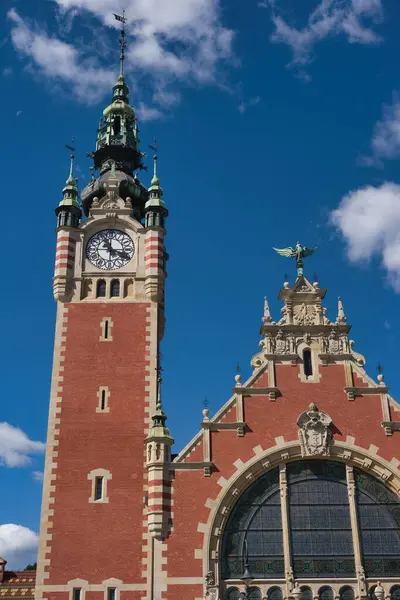 Uzun bir saat kulesi parlak mavi gökyüzünün altında detaylı mimarisi ile görkemli bir şekilde duruyor. Yapı, şehrin zengin tarihi ve kültürünü yansıtan dekoratif unsurlar sergiliyor..