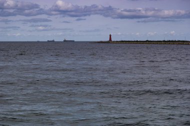 Sakin suların sakin manzarası ufka doğru uzanıyor. İskelenin ucunda bir deniz feneri duruyor. Bulutlu bir gökyüzünün altında çeşitli kargo gemileri görülebilir..