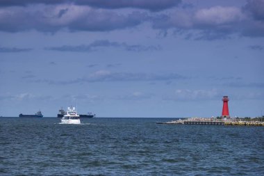 İki kargo gemisi uzaklarda yüzerken bir yat canlı kırmızı bir deniz fenerinin yanındaki sakin sularda geziniyor. Bulutlu gökyüzü kıyı sahnesi için sakin bir zemin sağlar..