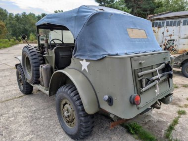 Eski bir askeri jip bulutlu bir gökyüzünün altında beton bir zeminde duruyor. Araç yumuşak bir üst kapağa sahip ve tarihi değerini vurgulayan yeşilliklerle çevrili..