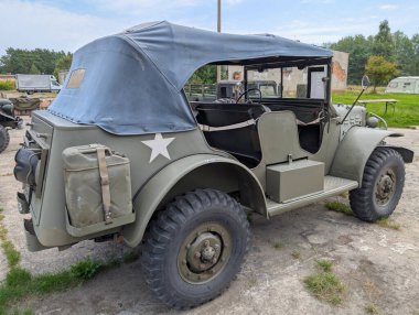 Mavi tuval kaplı klasik bir askeri araç, yeşil ve tarihi yapılarla çevrili çakıl taşlarının üzerine park edilmiştir..