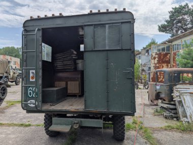 Eski bir askeri nakliye kamyonu engebeli bir sanayi bölgesinde duruyor. Arkası açık, ahşap sandıkları ve mobilyaları gösteriyor. Çevrede yıpranmış yapılar ve makineler var..