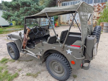 Açık havada klasik bir askeri jip sergilenirken, zeytin yeşili renkler ve klasik ayrıntılar yer alıyor. Çevrede kırsal bir cazibe var. Arka planda eski yapılar var..