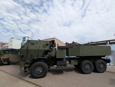 Büyük bir askeri taşıma aracı arka planda çeşitli binalar bulunan bir şehir caddesinde duruyor..