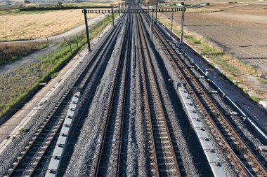 Demiryolu rayları düz ve kırsal bir araziye uzanır, temiz bir gökyüzünün altında. Çakıl yatakları rayları desteklerken, çevresindeki tarlalar gün ışığında sakin bir kırsal alanı gösteriyor..