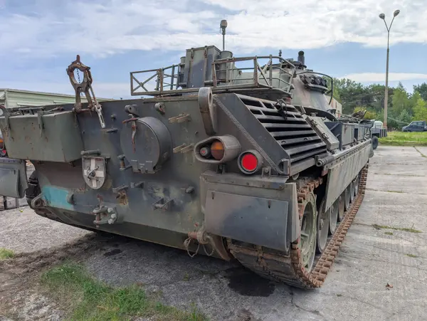 Bir askeri tank açık mavi gökyüzünün altındaki bir araç parkında park halinde. Tank kullanım belirtileri gösteriyor ve yeşil ağaçlarla çevrili. Bu da bakım için güvenli bir yer olduğunu gösteriyor..