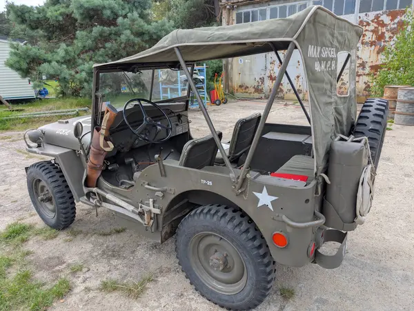 Açık havada klasik bir askeri jip sergilenirken, zeytin yeşili renkler ve klasik ayrıntılar yer alıyor. Çevrede kırsal bir cazibe var. Arka planda eski yapılar var..