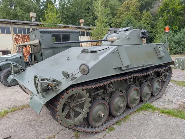 Takip edilmiş bir zırhlı personel taşıyıcı ağaçlarla çevrili eski bir askeri binanın önünde belirgin bir şekilde duruyor. Araç monte edilmiş bir silah ve belirgin bir kamuflaj içeriyor..