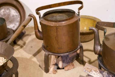 Tarihi bir mutfağı tasvir eden eski bir pirinç çaydanlık, diğer eski mutfak malzemelerinin yanında bir ahşap tezgahta duruyor. Atmosfer yemek pişirme geleneklerini ortaya çıkarıyor..