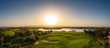 Şafak vakti, sakin bir göl, canlı bir yeşillik ve El Gouna Kızıldeniz 'deki ufuk çizgisinde parlayan gündoğumuyla yükselen sakin bir atmosfer içeren bir golf sahasının resimsel görüntüsü.