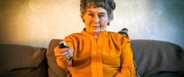 Sarı günlük kıyafetli yaşlı bir kadın evdeki rahat bir koltuğa oturur ve televizyon izler, büyükanne uzaktan kumandayı tutar ve kanalı değiştirir, haber veya TV dizisi izler..