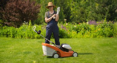 A young man in a straw hat is putting on gloves to mowe a lawn with a lawn mower in his beautiful green floral summer garden. A professional gardener with a lawnmower cares for the grass in the