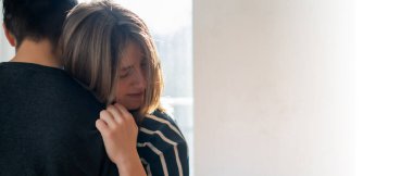 A young girl warmly and tenderly hugs her boyfriend, a couple stands against the background of a window on a sunny day, a woman begins romantic relationship. Husband and wife feel united, in harmony.