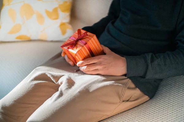 一个年轻人坐在沙发上 手里拿着一个小礼物 包装在盒子里 用带子绑在一个私密的地方 — 图库照片