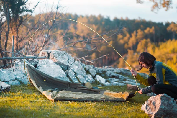 A young girl sets up her tent at a campsite on a green meadow among the mountains on a warm summer evening. The traveler is getting ready for a comfortable sleep in nature.