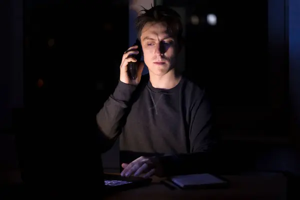 A young man types on a laptop keyboard, a freelancer works at night in a dark room, a student learns programming, plays computer games.
