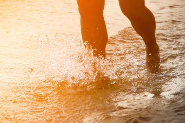 Suyun üzerinde sahil boyunca koşan biri, güneş ışınlarında okyanus ya da deniz, sporcu, antrenman yapıyor, dışarıda koşuyor, bacaklara yakından bakıyor ve su sıçratıyor..