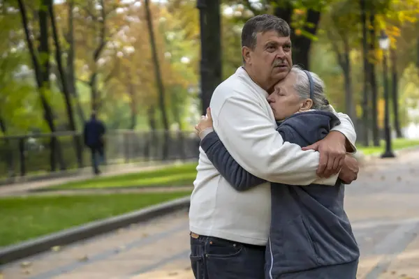 An elderly couple hugs in a park on a walk, a husband hugs his wife tightly and tenderly, supports her during her treatment against disease, a caring man and woman help each other in difficult times.