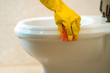 Sarı lastik eldivenli kadın eller bir bez tutuyorlar ve deterjan ve dezenfektanla tuvaleti siliyorlar. Kadın banyoyu temizliyor..