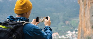Young man with a backpack and hiking equipment walks in the mountains and takes a photo on the mobile phone of a beautiful picturesque village. Traveler enjoys the scenery and shares photos.