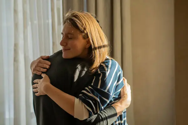 stock image Happy young girl warmly hugs her boyfriend, rejoices at the meeting, stands near the window, smiley face in close-up, woman starts a romantic relationship. Husband and wife feel united, in a harmony.