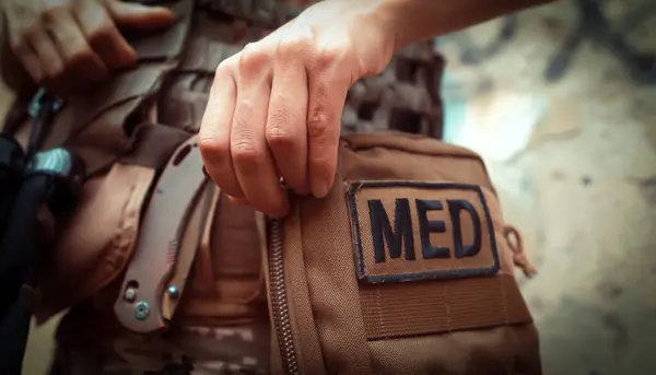 Kurşungeçirmez yelek giyen bir doktor, kamuflajlı, ilk yardım çantasını açar, bir kadın taktik tıp ile meşgul olur ve cephede yakın plan bir cephede hayat kurtarır..