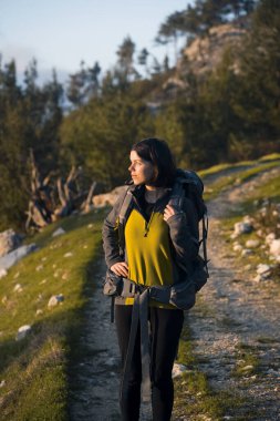 Sırt çantası ve seyahat teçhizatıyla genç bir kız, patika boyunca yürürken dağlardaki muhteşem günbatımını seyrediyor. Bir gezgin ulusal parkta yürüyor..