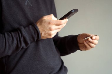 Male hands are holding a mobile phone and a bank card, a young man in a dark casual sweater pays for purchases, makes online banking using an application, close-up view.