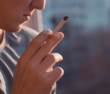 Genç bir adam sigara içiyor, elinde bir sigara tutuyor ve sabahın erken saatlerinde pencerenin önünde dikilirken dumanı içine çekiyor..