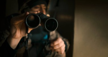 Askeri üniformalı ve kask takan genç adam savaş sırasında dürbünle bakıyor ve askeri tatbikatlara katılıyor. Asker binanın içerisindeki pencerenin yanında keşif yapıyor..