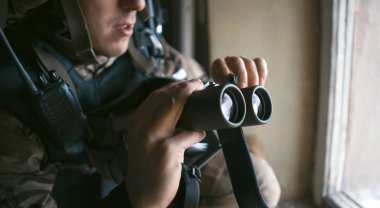 Askeri üniformalı ve kask takan genç adam savaş sırasında dürbünle bakıyor ve askeri tatbikatlara katılıyor. Asker binanın içerisindeki pencerenin yanında keşif yapıyor..