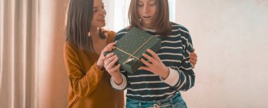 Two happy young girls laugh, friends hug and give each other gifts during the holiday. Women surprise and congratulate on the anniversary of their relationship in their cozy home.