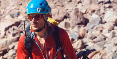 Güneş gözlüklü, kasklı ve kırmızı termal ceketli genç bir adam dağlarda tek başına yürüyor. Gezgin kayalara, taşlara tırmanıyor, dağcılık için içeri giriyor..